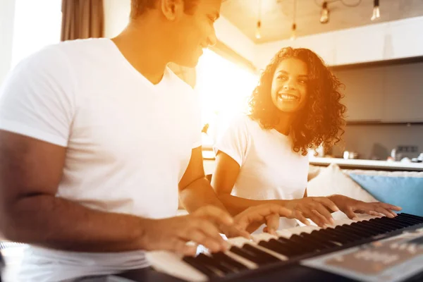 A black man sits in the living room of his apartment and plays a synthesizer. A girl is sitting next to him. — Stock Photo, Image