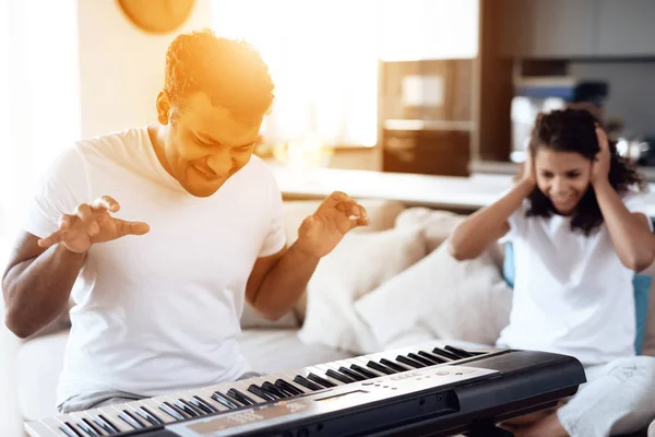 A black man sits in the living room of his apartment and plays a synthesizer. A girl sits next to him, covering her ears — Stock Photo, Image