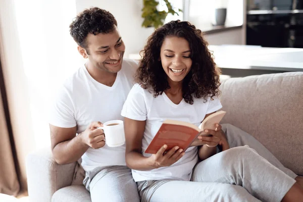 Черные мужчина и женщина сидят на диване. Женщина читает книгу, мужчина сидит и обнимает ее . — стоковое фото