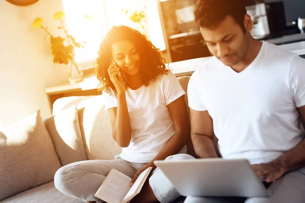 Black man and woman are sitting on the couch. A woman is reading a book, a man is sitting and working behind a laptop.