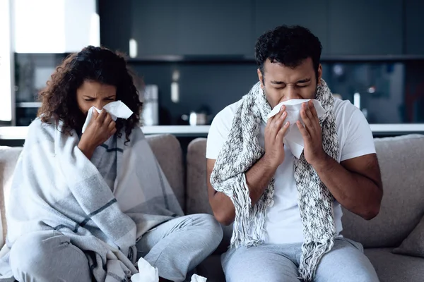 Hombres y mujeres negros están sentados en el sofá. Cogen un resfriado y se soplan la nariz en servilletas de papel . —  Fotos de Stock