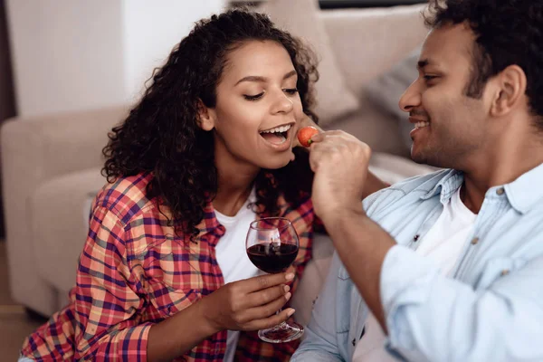 Svart man och kvinna sitter på soffan. En man och en kvinna omfamnade och drack vin och äta jordgubbar. — Stockfoto