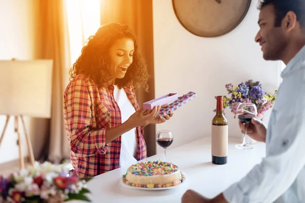 Black man and woman in the kitchen at home. The man has prepared a gift for the woman, which he hands her.