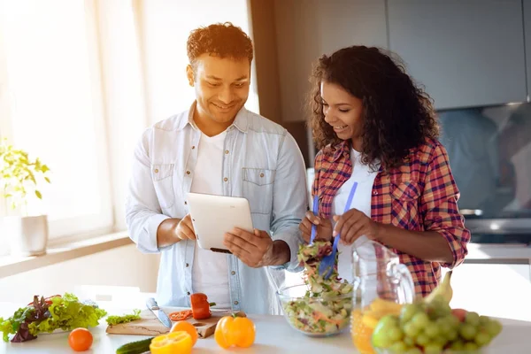 Svart man och kvinna i köket hemma. De förbereder frukost, mannen tittar på något på sin surfplatta. — Stockfoto