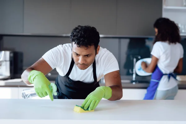 Hombres y mujeres negros en la cocina de casa. Limpian en la cocina. Un hombre lava la encimera con detergente . — Foto de Stock