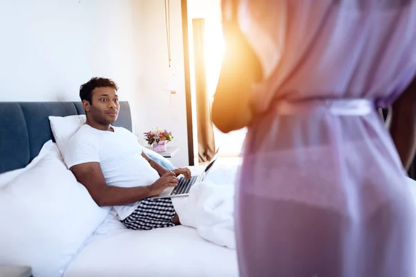 Black man and woman in the bedroom. A man is lying on the bed with laptop. Before him is his girl in sexy lingerie. — Stock Photo, Image