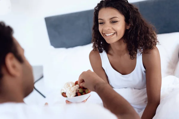 Hombres y mujeres negros se sientan en su cama por la mañana en el dormitorio. Un hombre está alimentando a una mujer una fresa con crema . —  Fotos de Stock