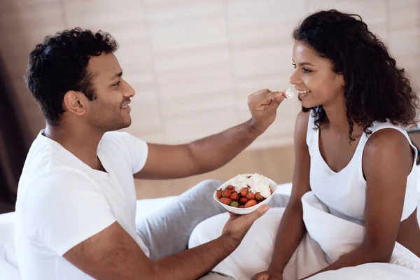 Black men and women sit in their bed in the morning in the bedroom. A man is feeding a woman a strawberry with cream. — Stock Photo, Image