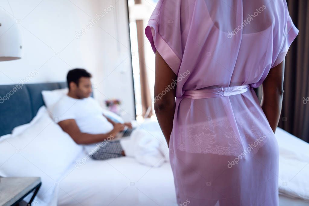 Black man and woman in the bedroom. A man is lying on the bed with laptop. Before him is his girl in sexy lingerie.