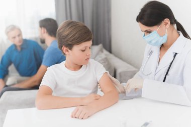 Hemşire insülin enjeksiyonu için çocuk yapar. Bir hemşire bir maske ve eldiven masada oturuyor. Çocuğun evde olduklarını. Yaşlı bir adam ve bir adam arkasında oturmak.