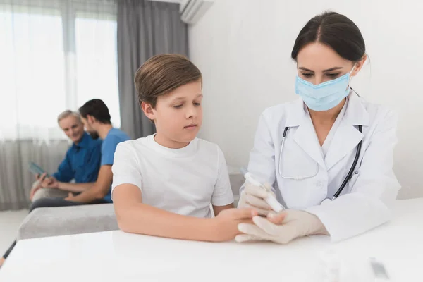 Eine Krankenschwester macht einen Bluttest mit dem Vertikutierer eines Jungen. eine Krankenschwester sitzt mit Maske und Handschuhen am Tisch. — Stockfoto