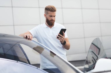 Onun elektrikli otomobil yakınındaki telefonda konuşurken sakallı genç bir adam.