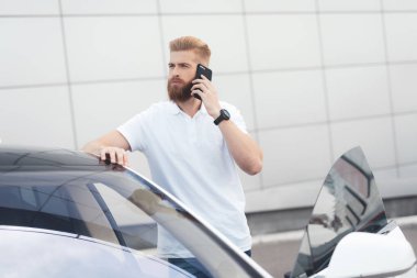 Onun elektrikli otomobil yakınındaki telefonda konuşurken sakallı genç bir adam.