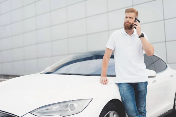 髭の彼の電気自動車近く電話で話している若い男. — ストック写真