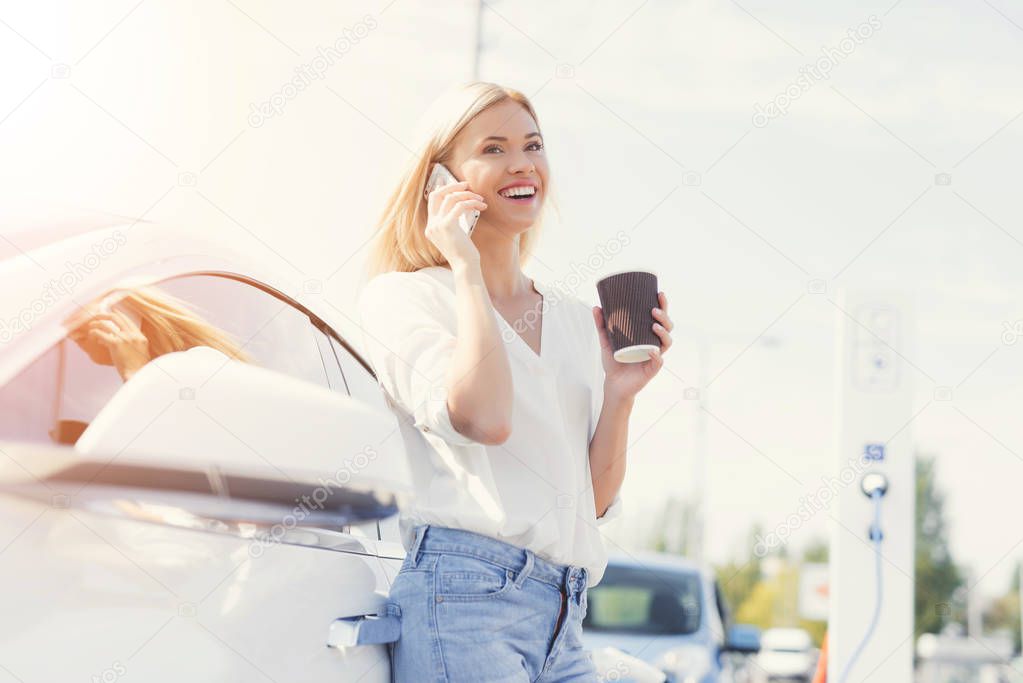 A blonde girl drinks coffee and talks on the phone at an charging station.