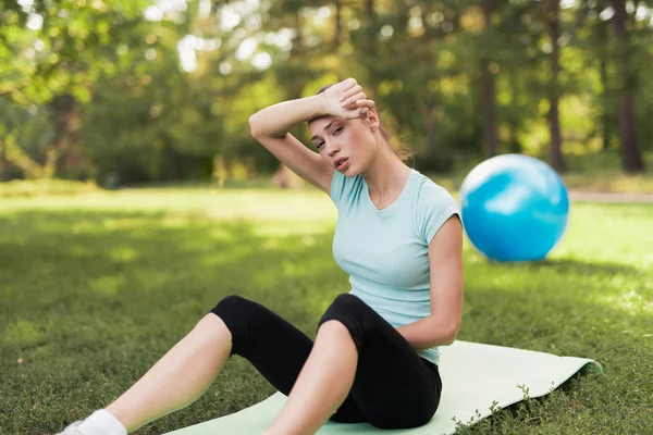 Una mujer está sentada en una alfombra para el yoga y descansando después de un entrenamiento. Detrás de ella hay una bola azul para el yoga . —  Fotos de Stock