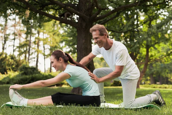 Чоловік допомагає жінці розтягуватися. Вона сидить на килимку для йоги — стокове фото