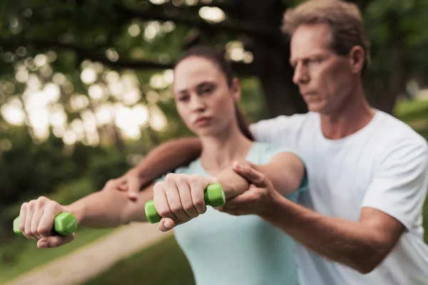 Girl doing exercises with dumbbells in the park. A man helps her. They are smiling — Stock Photo, Image
