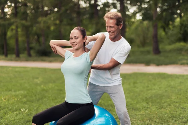 Ein Mann trainiert eine Frau, die Übungen macht, während sie im Park auf einem Ball für Yoga sitzt — Stockfoto
