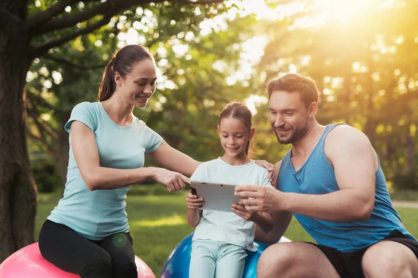 The family rests in the park after playing sports. They are looking at something on the tablet — Stock Photo, Image