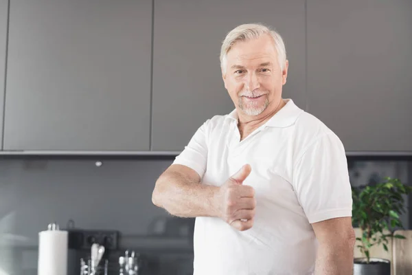 Quell'uomo è in cucina. Mostra il pollice in alto. Ha una maglietta bianca, una barba grigia e capelli . — Foto Stock
