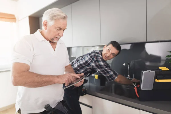Два водопроводчика стоят на кухне. Пожилой человек смотрит что-то на планшете, пока молодой человек чинит кран. . — стоковое фото