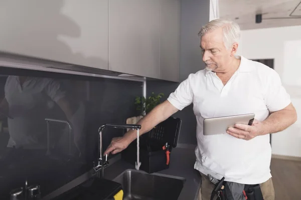 灰色の配管は、水道の蛇口の横にある台所に立っています。彼はクレーンの作業をチェックし、彼の手で灰色のタブレットを保持しています。 — ストック写真
