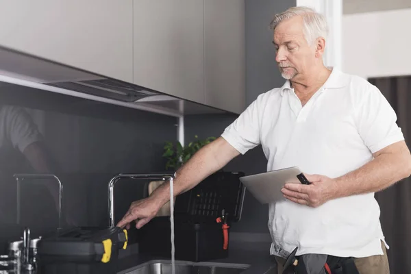 Šedé instalatér stojí v kuchyni u kohoutku. Kontroluje práci jeřábu a má šedé tabletu ve svých rukou — Stock fotografie