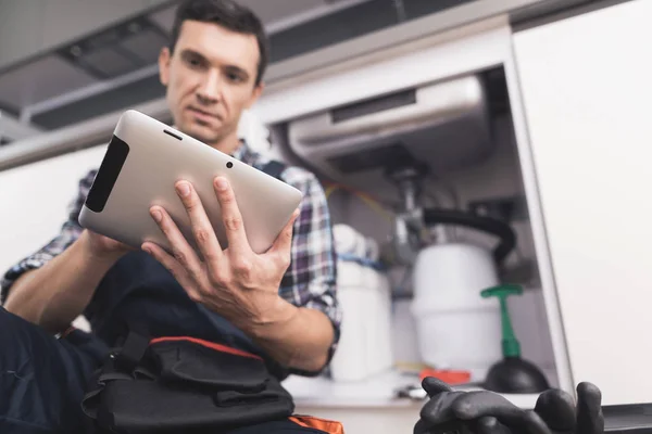 De loodgieter zit naast het aanrecht op de vloer en kijkt naar de tablet voor reparatie instructies. — Stockfoto