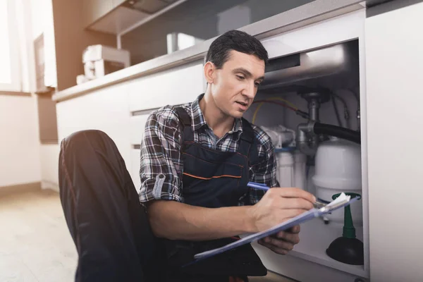 The plumber sits next to the kitchen sink on the floor and writes a report on the work done. — Stock Photo, Image