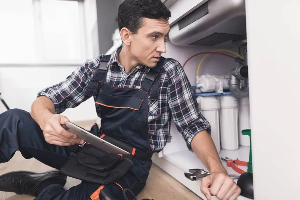 The plumber sits next to the kitchen sink on the floor and looks at the tablet for repair instructions. — Stock Photo, Image