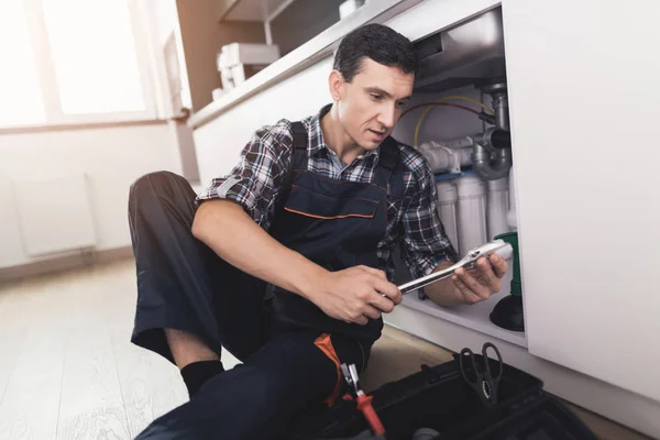 Ο υδραυλικός κάθεται δίπλα στο νεροχύτη της κουζίνας στο πάτωμα και επιλέγει το κατάλληλο κλειδί για την επισκευή του σωλήνα. — Φωτογραφία Αρχείου