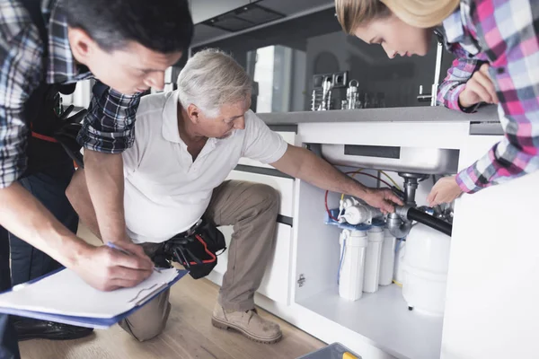 Una mujer le muestra a los plomeros un fregadero de cocina roto. Uno de los hombres inspecciona las tuberías, en segundo lugar escribe información al formulario . — Foto de Stock