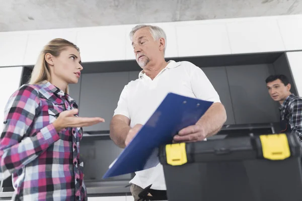 The woman is shocked by the bill for repairs from plumbers. An elderly man holds a blank in his hands. A young plumber examines the kitchen sink. A black toolbox is next to it.