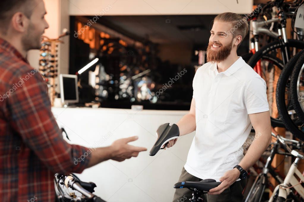 A seller at a bicycle store helps a young buyer choose a saddle for a bicycle. A man with a beard shows the client different types of goods. They both have a good mood.