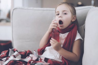 Küçük hasta kız kırmızı bir fular sarılı beyaz bir kanepede oturuyor. Boğazını tıbbi bir sprey ile su sıçramalarına