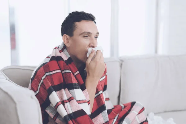 En man med en kall sitter på soffan, gömmer sig bakom en röd matta. Han blåser näsan i en servett — Stockfoto