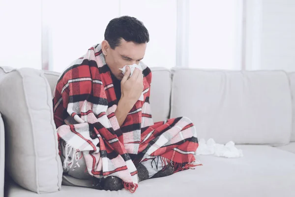 A man with a cold sits on the couch, hiding behind a red rug. He blows his nose into a napkin — Stock Photo, Image