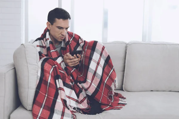 Человек с простудой сидит на диване, прячась за красным ковром. Он смотрит на свой смартфон — стоковое фото