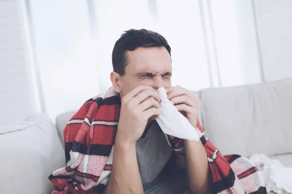 一个感冒的人坐在沙发上, 躲在红地毯后面。他把鼻子吹进餐巾 — 图库照片