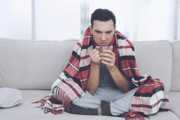 Człowiek z zimna siedzi na kanapie, chowając się za czerwony dywan. Pije herbaty lecznicze — Zdjęcie stockowe
