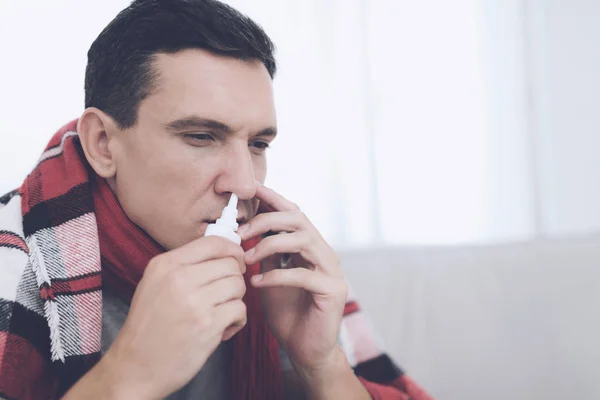 Un homme avec un rhume est assis sur le canapé, caché derrière un tapis rouge. Il asperge son nez de vaporisateur nasal — Photo