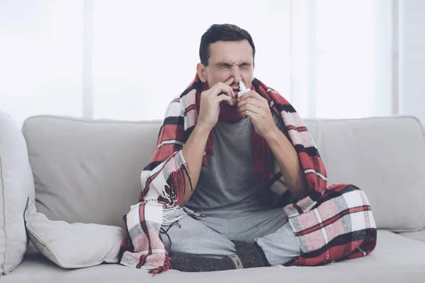 Człowiek z zimna siedzi na kanapie, chowając się za czerwony dywan. Kropi aerozol do nosa — Zdjęcie stockowe