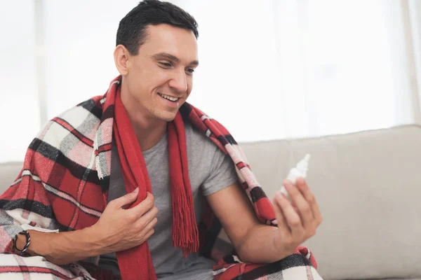 A man with a cold sits on the couch, hiding behind a red rug. He sprinkles nasal spray into his nose — Stock Photo, Image