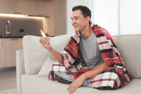 Un hombre con un resfriado se sienta en el sofá, escondido detrás de una alfombra roja. Él espolvorea aerosol nasal en su nariz — Foto de Stock