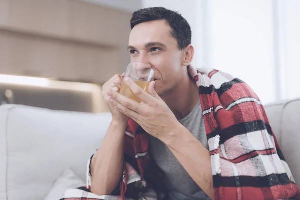 Un hombre con un resfriado se sienta en el sofá, escondido detrás de una alfombra roja. Bebe té medicinal. — Foto de Stock
