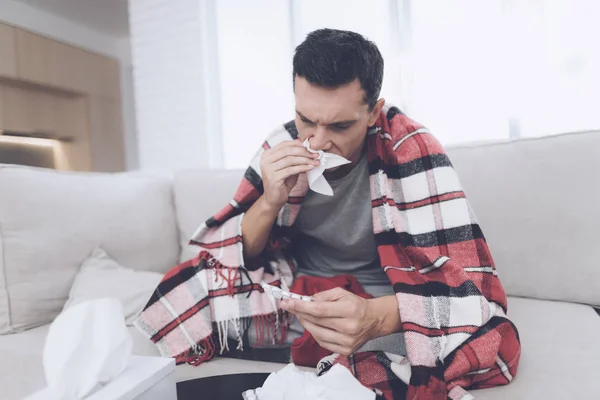 A man with a cold sits on the couch, hiding behind a red rug. Before him a lot of medicines — Stock Photo, Image