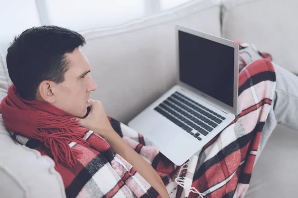 Ένας άνθρωπος με ένα κρύο κάθεται στον καναπέ, κρύβονται πίσω από ένα κόκκινο χαλί. Κάθεται με το laptop του στην αγκαλιά του — Φωτογραφία Αρχείου