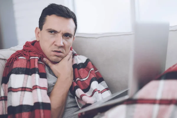 Człowiek z zimna siedzi na kanapie, chowając się za czerwony dywan. Siedzi z laptopem na kolanach — Zdjęcie stockowe