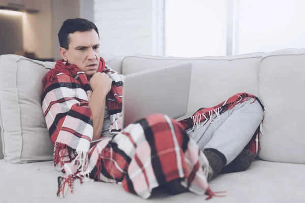 A man with a cold sits on the couch, hiding behind a red rug. He is sitting with his laptop on his lap